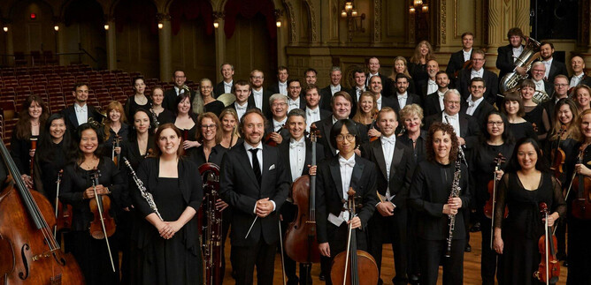 A large orchestra community is standing on stage. All musicians are elegantly dressed and smiling for the photo. | © VANC