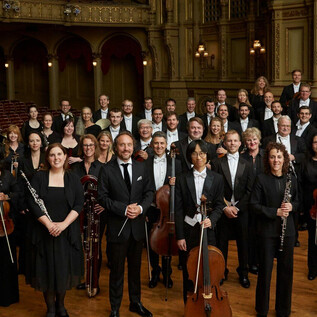 A large orchestra community is standing on stage. All musicians are elegantly dressed and smiling for the photo. | © VANC