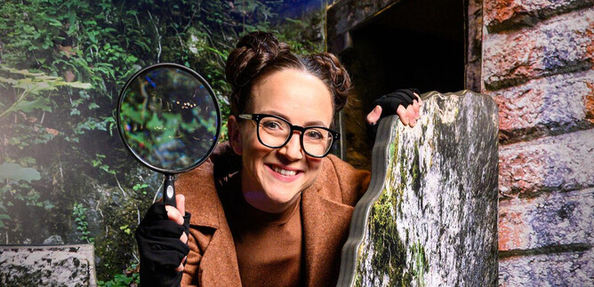 A cheerful woman with a magnifying glass stands in front of a rocky environment. She is wearing glasses and has two bun hairstyles. | © SLT / Tobias Witzgall