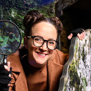 A cheerful woman with a magnifying glass stands in front of a rocky environment. She is wearing glasses and has two bun hairstyles. | © SLT / Tobias Witzgall