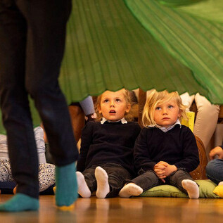 Two small children sit captivated on the floor, watching a dancer. Around them is a cheerful atmosphere with colorful cushions and other children. | © Erika Mayer