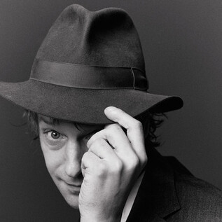 Muž nosí klobouk a usmívá se do strany. Záběr je v černobílé. | © Susanne Hassler-Smith