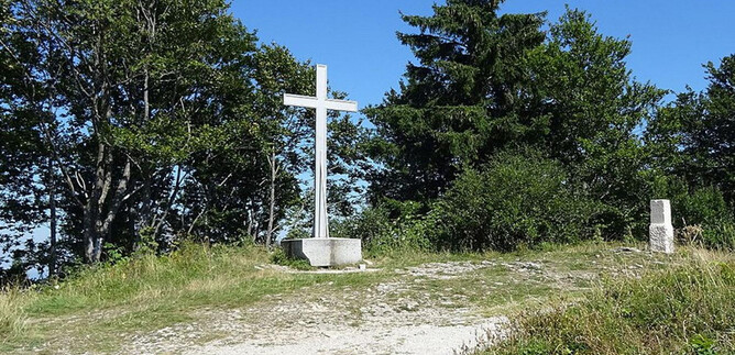 A white cross stands on a path, surrounded by trees and shrubs. The clear blue sky gives the scene a peaceful atmosphere. | © Benjamin Huber