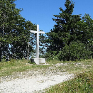 A white cross stands on a path, surrounded by trees and shrubs. The clear blue sky gives the scene a peaceful atmosphere. | © Benjamin Huber