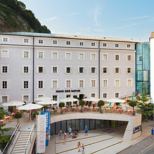 A modern building with a bright facade and glass accents. In front of the building, there are terraces with seating and plants. | © HdN_Simmerstatter