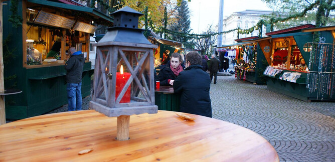 A cozy Christmas market with festively decorated stalls and people engaging in conversation. A lantern with red light stands in the foreground on a wooden table.