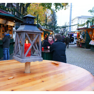 A cozy Christmas market with festively decorated stalls and people engaging in conversation. A lantern with red light stands in the foreground on a wooden table.