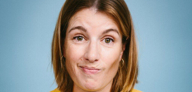 A woman with shoulder-length hair is wearing a yellow T-shirt. She has a neutral facial expression against a blue background. | © Apollonia Theresa Bitzan