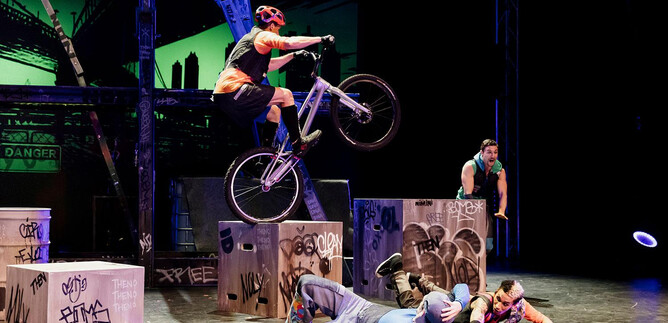 A cyclist jumps over boxes on a stage. In the background, other performers can be seen staging an action-packed scene in graffiti style. | © Caroline Thibault