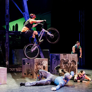 A cyclist jumps over boxes on a stage. In the background, other performers can be seen staging an action-packed scene in graffiti style. | © Caroline Thibault