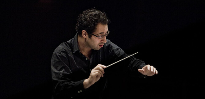 A conductor stands on stage and conducts with a baton. Behind him, a dark backdrop is visible. | © Pia Clodi
