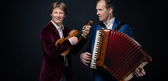 Two musicians stand against a dark background. One plays the violin, while the other holds the accordion. | © Severin Koller