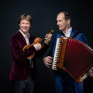 Two musicians stand against a dark background. One plays the violin, while the other holds the accordion. | © Severin Koller
