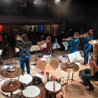 A band is playing on a stage with various instruments. The musicians are standing in a festive space with an enthusiastic crowd in the background. | © bodenst@ndig