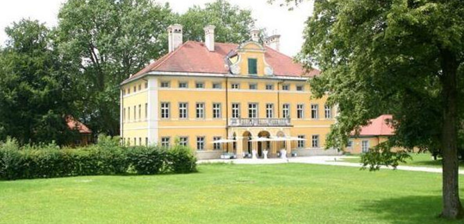 A large yellow building with a red roof stands in a green landscape. Surrounding the building are trees and a well-kept lawn. | © Christian Schneider