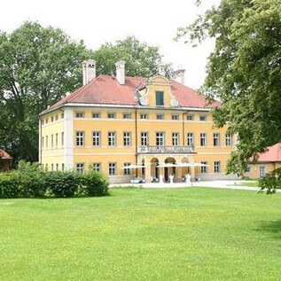A large yellow building with a red roof stands in a green landscape. Surrounding the building are trees and a well-kept lawn. | © Christian Schneider
