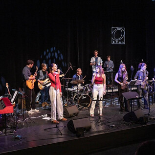 A band is performing on a stage, surrounded by instruments such as a piano, guitar, and drums. Two female singers are in the foreground singing into a microphone. | © Musikum Salzburg