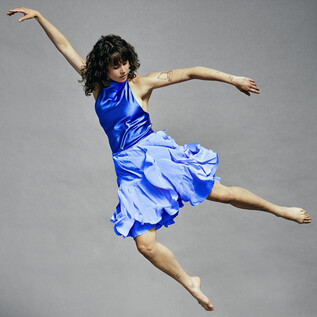 A dancer in a blue dress jumps dynamically through the air. The background is gray and neutral. | © Chris Rogl