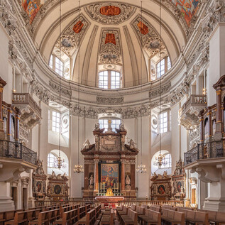 A magnificent church with a high dome and elaborate ceiling frescoes. Benches and organs flank the altar in the center. | © Salzburger Dom / Eva trifft. Fotografie