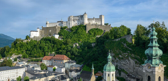An impressive castle on a hill, surrounded by green nature. In the foreground, the roofs of a city with historical buildings are visible. | © Tourismus Salzburg / G. Breitegger