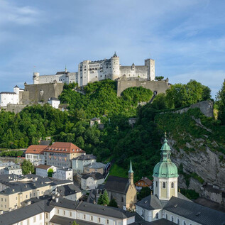 An impressive castle on a hill, surrounded by green nature. In the foreground, the roofs of a city with historical buildings are visible. | © Tourismus Salzburg / G. Breitegger