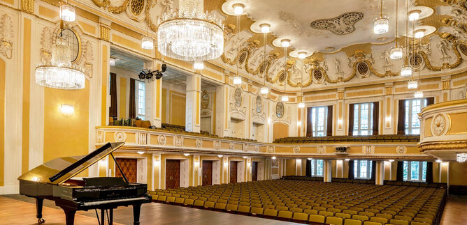 An elegant concert hall with a black piano on stage. The seated rows and the elaborate ceiling style give the room a festive atmosphere. | © Stiftung Mozarteum Salzburg / Andrew Phelps