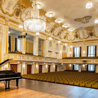 An elegant concert hall with a black piano on stage. The seated rows and the elaborate ceiling style give the room a festive atmosphere. | © Stiftung Mozarteum Salzburg / Andrew Phelps