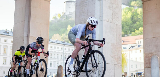 Three cyclists are riding under an arch. The landscape in the background shows a city with green hills and historic buildings. | © Cyclodom