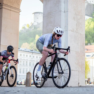 Three cyclists are riding under an arch. The landscape in the background shows a city with green hills and historic buildings. | © Cyclodom